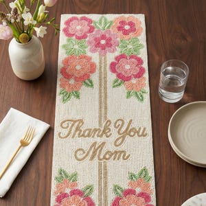 Puede incluir: Camino de mesa con cuentas con diseños florales en rosa, coral y verde. El camino de mesa presenta el texto "Thank You Mom" en cuentas doradas. Un jarrón con flores, un vaso de agua y un plato están sobre la mesa de madera.