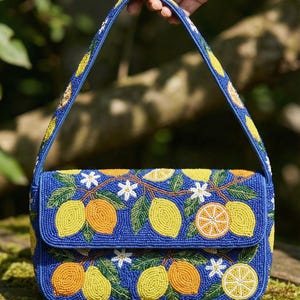 May include: A blue beaded handbag with a flap closure and a handle. The bag is decorated with yellow lemons, orange citrus fruits, white flowers, and green leaves. The handle and edges of the bag are also beaded with yellow and blue beads.