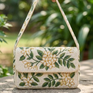 May include: Cream-colored beaded handbag with a flap closure, adorned with floral and leaf patterns in green and yellow. The bag has a shoulder strap also decorated with beads. The background is blurred, suggesting an outdoor setting.