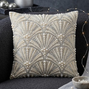 Puede incluir: Cojín decorativo cuadrado con un diseño en forma de abanico hecho de cuentas y perlas de imitación de color blanco roto y plateado. El cojín está colocado sobre una superficie gris oscuro.