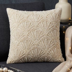 Può includere: Cuscino decorativo quadrato color crema con un motivo a ventaglio di perline. Il cuscino è posto su un divano grigio scuro. Il design a perline crea una superficie strutturata, aggiungendo un tocco di eleganza all'arredamento.