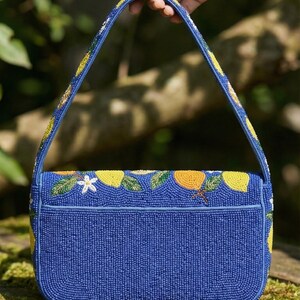May include: A blue beaded handbag with a rectangular shape and a top handle. The bag is decorated with yellow lemons, white flowers, and green leaves. The handle and edges are also adorned with yellow and white beads.