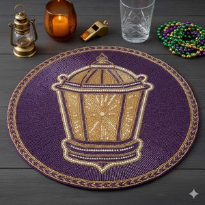 Op de afbeelding: Een ronde, paarse placemat met kralen en een gouden lantaarnontwerp in het midden. De lantaarn is gedetailleerd met gouden en witte kralen. Een gouden kralenrand omringt de placemat. Decoratieve items zoals een kaars, fluitje en kralen staan op de achtergrond.