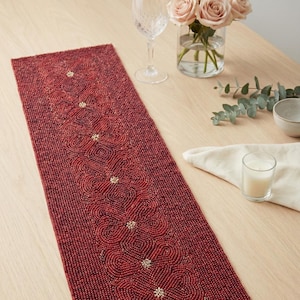Może przedstawiać: Czerwony, koralikowy bieżnik na stół z kwiatowym wzorem. Bieżnik jest prostokątny i posiada powtarzający się wzór kwiatów z małymi, złotymi akcentami. Bieżnik leży na jasnym drewnianym stole.
