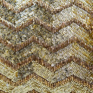 K&ouml;nnte beinhalten: Nahaufnahme eines gold- und braunen Stoffes mit Chevron-Muster. Der Stoff besteht aus Pailletten und Perlen.