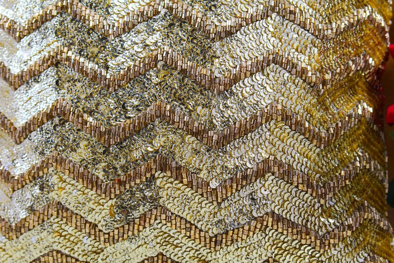 K&ouml;nnte beinhalten: Nahaufnahme eines gold- und braunen Stoffes mit Chevron-Muster. Der Stoff besteht aus Pailletten und Perlen.