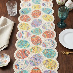 May include: Beaded Easter egg table runner in cream, with colorful eggs in shades of blue, yellow, orange, and purple. The runner is on a dark wood table, with a glass, plate, and flowers.