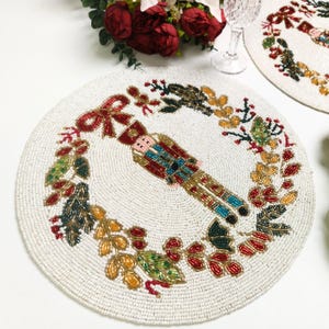 May include: Round, beaded placemat with a festive design. The off-white mat features a detailed image of a nutcracker soldier surrounded by a wreath of colorful leaves and berries. A red bow is at the top. The mat is likely used for holiday or Christmas decor.