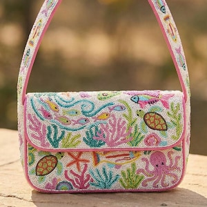 Könnte beinhalten: Eine weiße, perlenbesetzte Handtasche mit rosa Riemen und Besatz. Die Tasche zeigt eine Unterwasserszene mit bunten Fischen, Korallen, Meeresschildkröten und einem Oktopus. Das Design ist detailliert und lebendig.