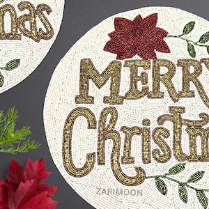May include: White beaded round placemat with the words "Merry Christmas" in gold beads. A red beaded poinsettia flower and green beaded leaves are on the placemat.