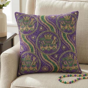 May include: Square purple pillow with a beaded design featuring green, gold, and purple accents. The pillow has a repeating pattern of circular designs with a mask, crown, and musical instruments. A beaded necklace with green, gold, and purple beads is in the foreground.