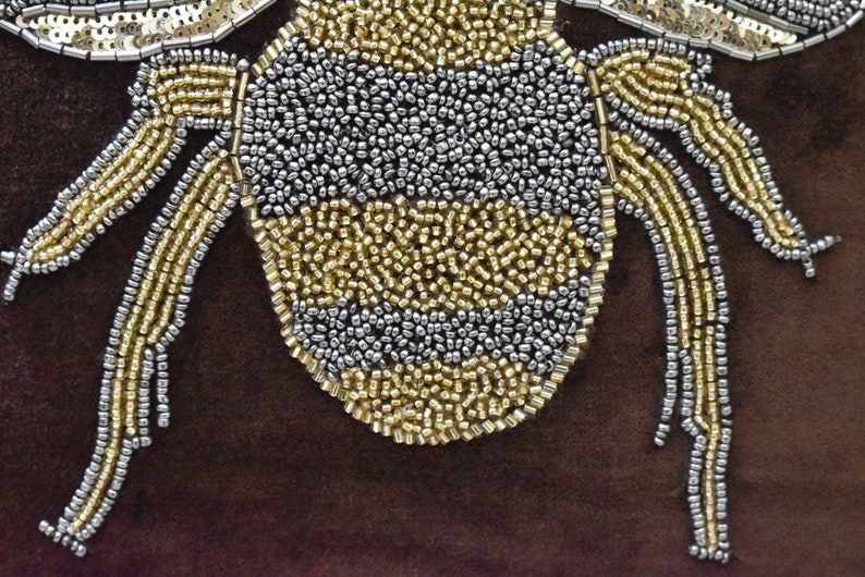 May include: A close-up of a beaded bumble bee with a gold and silver design. The bee is on a brown background.