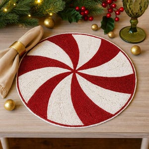 May include: A round beaded placemat with a red and white spiral design. The placemat is made of small beads and has a candy cane appearance. The placemat is on a wooden table, with a napkin and gold napkin ring, and Christmas decorations in the background.