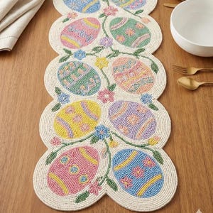 以下が含まれることがあります： ピンク、青、黄色、緑色のカラフルなイースターエッグのデザインが施されたビーズのイースターテーブルランナー。卵は花の模様で装飾され、つるや花に囲まれています。ランナーは木製のテーブルの上にあります。