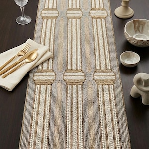 May include: Beaded table runner with a column design in neutral tones of beige, white, and gray. The runner is rectangular and features vertical stripes, creating a textured surface. Decorative elements like a candle, bowls, and silverware are on the table.