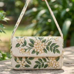 May include: Cream-colored beaded handbag with a floral design. The bag features a flap closure and a shoulder strap, both adorned with beads. The floral pattern includes yellow and green beaded flowers and leaves, creating a detailed and elegant accessory.