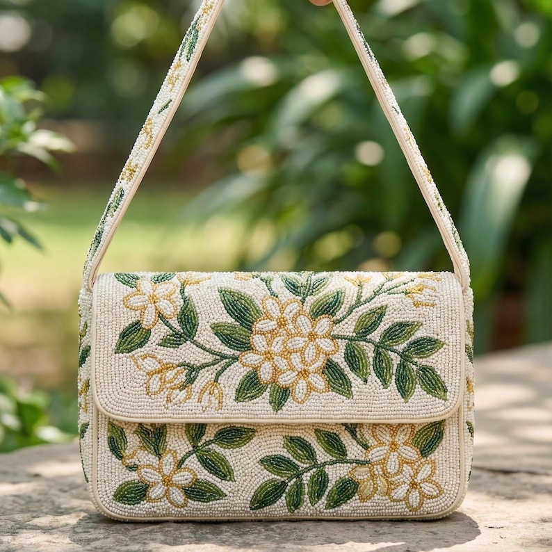 May include: Cream-colored beaded handbag with a floral design. The bag features a flap closure and a shoulder strap, both adorned with beads. The floral pattern includes yellow and green beaded flowers and leaves, creating a detailed and elegant accessory.