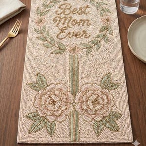 Puede incluir: Camino de mesa con cuentas con el texto "Best Mom Ever" en escritura dorada, rodeado por una corona floral. Presenta dos grandes diseños florales rosas en la parte inferior, con hojas verdes y un diseño de rayas verticales.