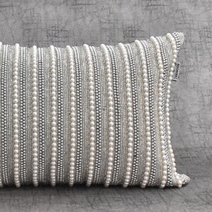 May include: A white and silver decorative pillow with a beaded design. The pillow has a label that says "ZAKKNOON".