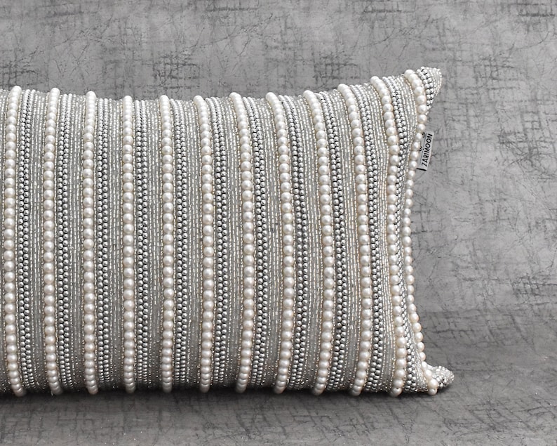 May include: A white and silver decorative pillow with a beaded design. The pillow has a label that says "ZAKKNOON".