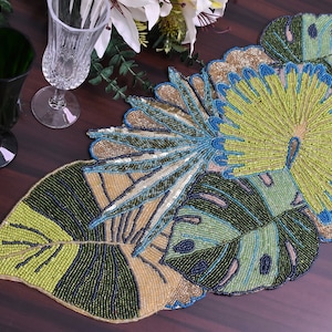 May include: A beaded table runner featuring a tropical leaf design. The runner is made from green, blue and beige beads and has a large, stylised leaf in the centre.