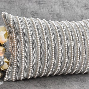 May include: A white and silver rectangular pillow with a beaded design. The pillow is covered in rows of small white beads and silver beads.