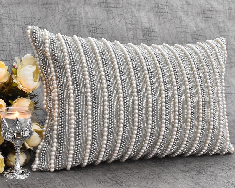 May include: A white and silver rectangular pillow with a beaded design. The pillow is covered in rows of small white beads and silver beads.