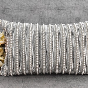 May include: A white and silver beaded lumbar pillow with a subtle sheen. The pillow is rectangular and has a decorative pattern of vertical rows of beads.