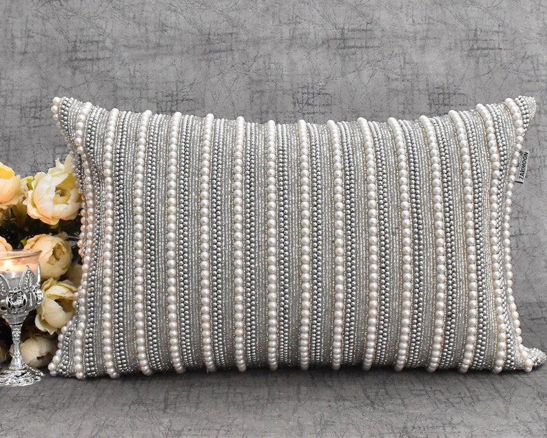 May include: A white and silver beaded lumbar pillow with a subtle sheen. The pillow is rectangular and has a decorative pattern of vertical rows of beads.