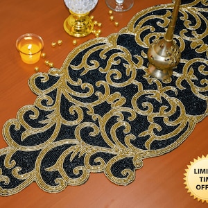 Op de afbeelding: Een zwarte en gouden kralen tafelkleed met een bloemmotief. De tafelkleed ligt op een houten tafel met een kaars en andere decoratieve items.