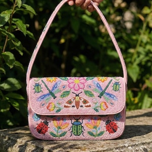 Op de afbeelding: Roze kralen handtas met een klep en handvat. De tas is versierd met kleurrijke kralenontwerpen van insecten, bloemen en bladeren. De achtergrond is een groene tuin.