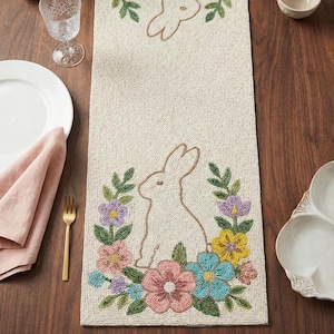 May include: Beige beaded Easter table runner with a bunny and floral design. The runner features a bunny outline surrounded by colorful flowers in pink, blue, yellow, and purple. The runner is rectangular.