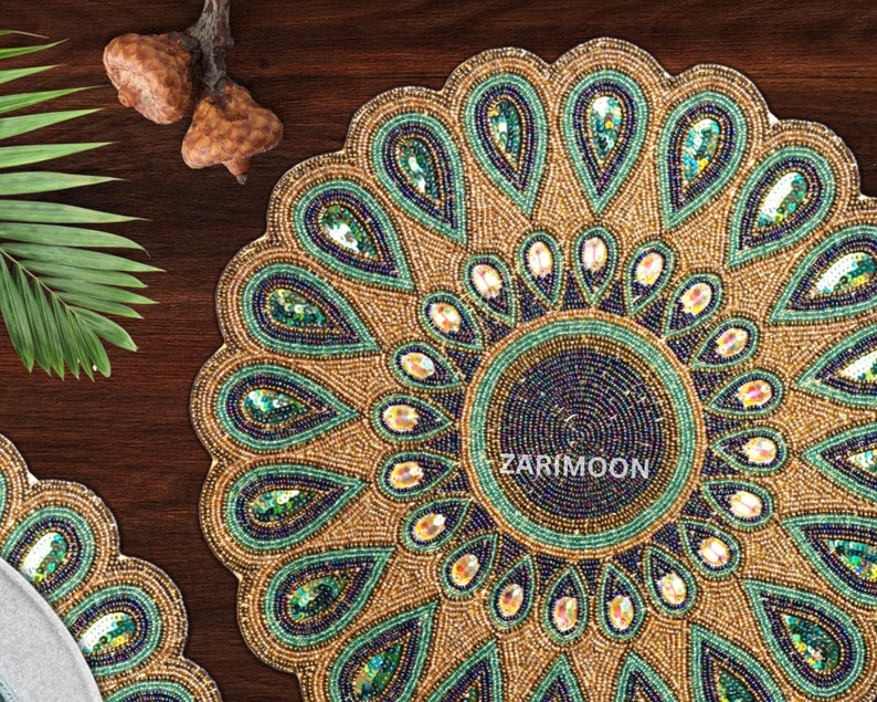May include: A round beaded table mat with a peacock feather design. The mat is gold with green and blue accents. The center of the mat is a dark blue circle with the text "ZARIMOON" in white.
