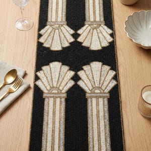 Può includere: Runner da tavolo nero con perline con un motivo ripetuto di colonne stilizzate e disegni a forma di ventaglio in crema, oro e argento. Il runner rettangolare è su una superficie di legno, con stoviglie e oggetti decorativi.