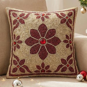 Op de afbeelding: Vierkant decoratief kussen met een bloemmotief. Het kussen heeft een beige achtergrond met een kralenpatroon. Het bloemmotief is in een dieprode kleur met rode edelsteenaccenten. Het kussen ligt op een beige bank.