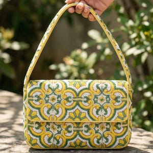 Peut inclure: Un sac à main rectangulaire en perles avec un motif floral jaune, vert et blanc. Le sac a une poignée et une bordure jaunes. Le sac est tenu par une main sur un fond vert flou.
