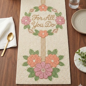 Puede incluir: Camino de mesa con cuentas y diseño floral. El camino de mesa blanquecino presenta una franja vertical central y una corona floral con el texto "For All You Do". También hay flores rosas y melocotón con hojas verdes.