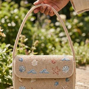 Peut inclure: Un sac à main en perles beige doté d'une fermeture à rabat et d'une anse unique. L'extérieur est orné de petites perles de verre colorées formant des motifs de coquillages, d'étoiles de mer et de vagues dans des tons bleu, blanc et pêche.
