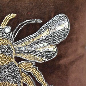 May include: A brown velvet pillow with a beaded bee design. The bee is made of gold and silver beads and has a shiny, iridescent finish.