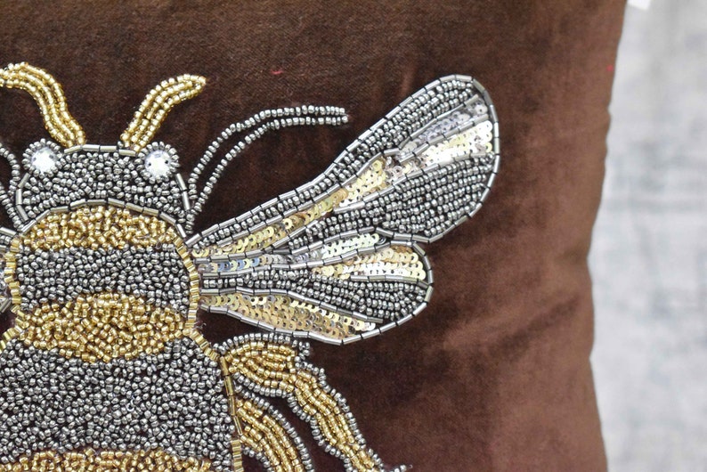 May include: A brown velvet pillow with a beaded bee design. The bee is made of gold and silver beads and has a shiny, iridescent finish.