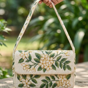May include: Cream-colored beaded handbag with a floral design. The bag features a flap closure and a beaded handle. The floral pattern includes white and yellow flowers with green leaves. The background is blurred.