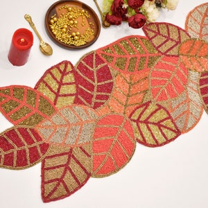 May include: A beaded table runner featuring a leaf design in shades of red, gold, and peach. The runner is crafted from small beads sewn together to create a textured, three-dimensional effect.