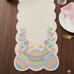 以下が含まれることがあります： ベージュのビーズイースターテーブルランナー、スカラップエッジ付き。ランナーは、パステルカラーのイースターエッグと、ピンク、黄色、紫色の花のアクセントが刺繍されています。濃い色の木製のテーブルの上に置かれています。