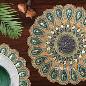 May include: Two round beaded placemats with a peacock feather design. The placemats are gold, green, and blue with a circular pattern in the center. The placemats are on a dark brown wooden surface.