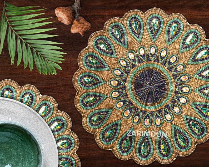 May include: Two round beaded placemats with a peacock feather design. The placemats are gold, green, and blue with a circular pattern in the center. The placemats are on a dark brown wooden surface.