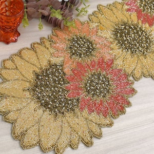May include: A beaded table runner with a sunflower design. The runner is made of gold, peach, and coral beads.
