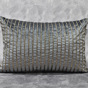 Puede incluir: Una almohada gris y dorada a rayas con un brillo sutil. La almohada está hecha de una tela suave, similar al terciopelo, y presenta un delicado patrón de cuentas doradas.