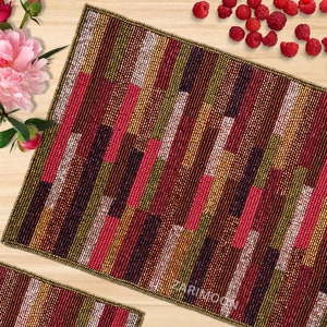 Puede incluir: Un mantel individual rectangular de cuentas con un patrón geométrico en tonos de rojo, marrón, dorado y blanco. El mantel individual está hecho de pequeñas cuentas tejidas juntas para crear una superficie texturizada.