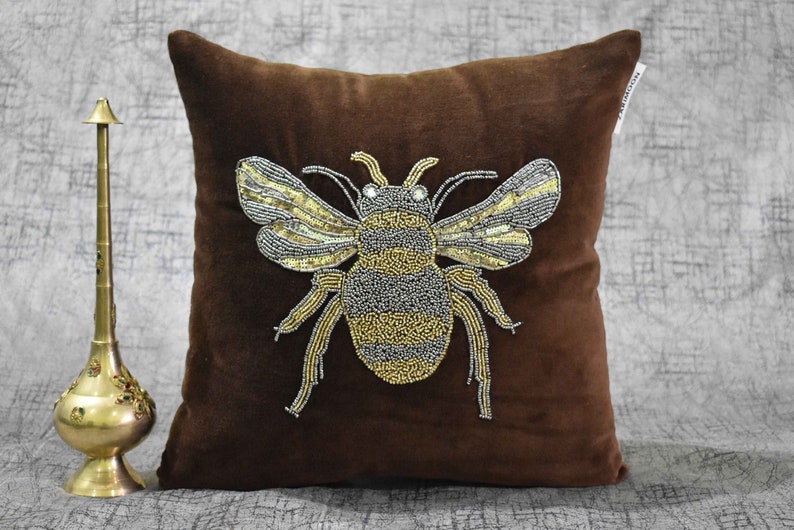 May include: A brown velvet pillow with a beaded bee design. The bee is made of gold and silver beads and has a detailed design.