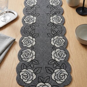 May include: A grey beaded table runner with a scalloped edge, featuring white and grey rose designs. The runner is displayed on a wooden table, showcasing a decorative floral pattern. This elegant table runner is perfect for special occasions.